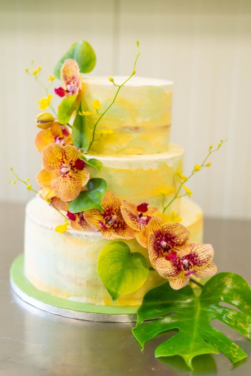 Een wit en gele bruidstaart in drie lagen met sierlijke bloemen en groene bladeren.