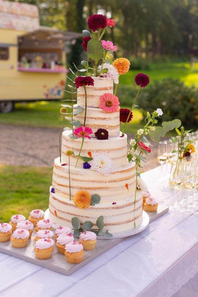 Een hoge bruidstaart met strakke afwerking en kleurrijke bloemen. Naast de taart liggen kleine muffins.